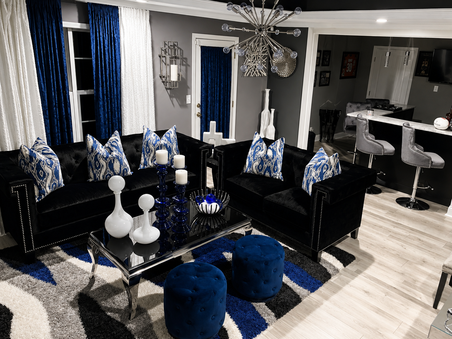 Living room with black tufted sofas, royal blue ottomans, chrome sputnik chandelier, and abstract wave rug