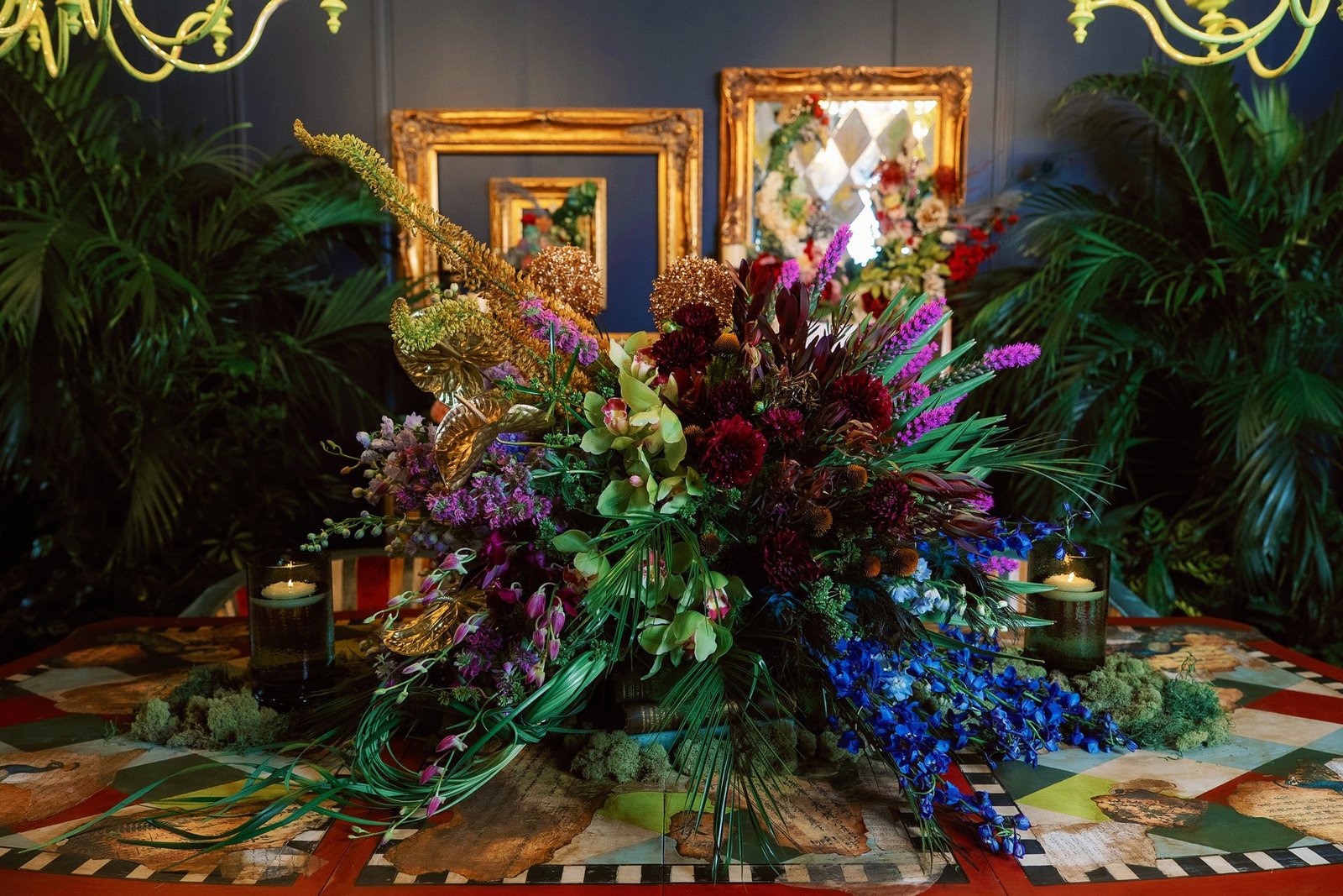 Styled dining table with a lush floral centerpiece of purple dahlias, blue delphiniums, and gilded leaves flanked by green glass candles
